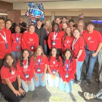 group of students wearing red shirts posing in an airport terminal with a patriotic mylar balloon