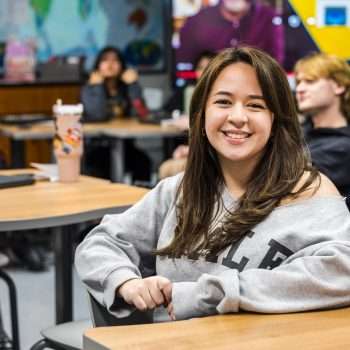 female high school student sitting at table inside of a classroom wearing a grey sweatshirt