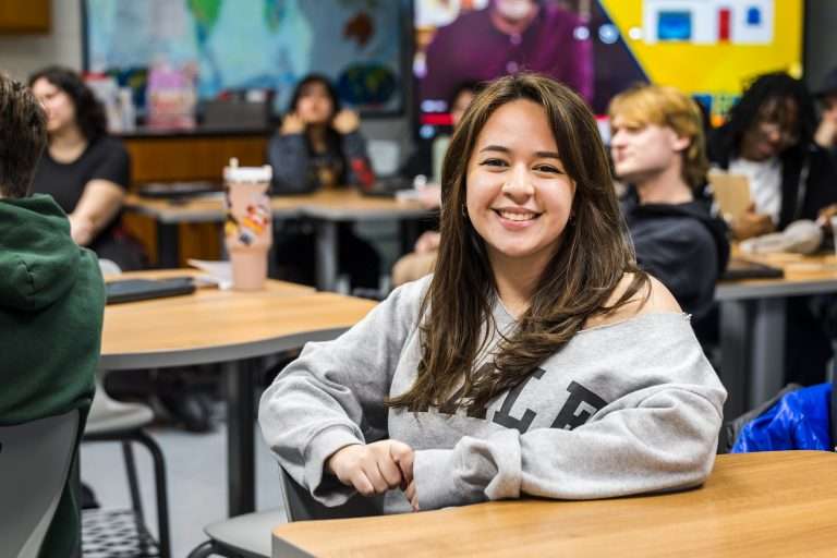 female high school student sitting at table inside of a classroom wearing a grey sweatshirt