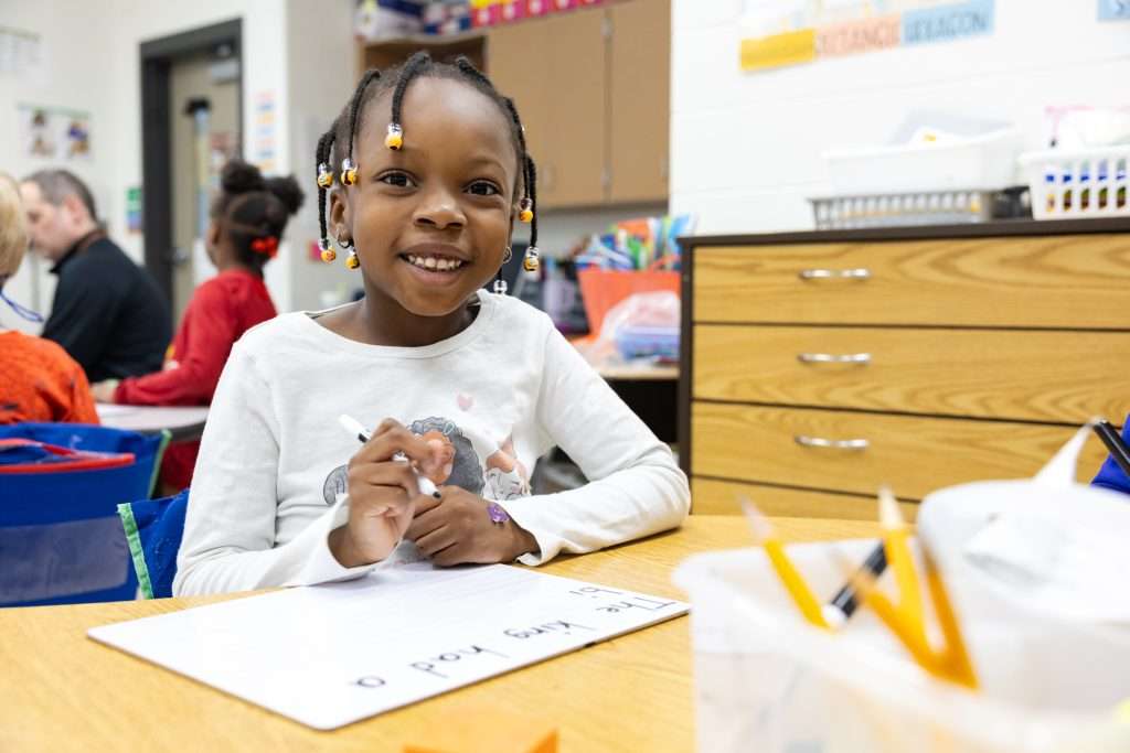 female elementary student sitting at table smiling with beaded hair writing on a white board