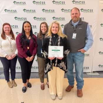 Group of five adults, four female and one male, standing in front of a CMCSS backdrop. Female with blond hair wearing a black sweater and cream colored pants is holding a certificate.