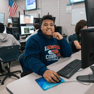 Boy wearing blue sweatshirt smiles at camera behind computer screen.