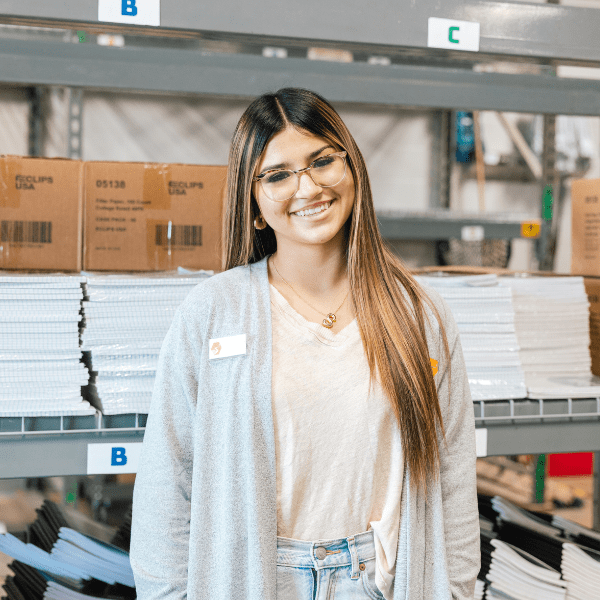 Teen with glasses and long hair standing in a warehouse with metal racks behind her.