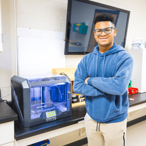 Teen in blue sweatshirt has arms folded in front of chest, standing at a 3D printer on the table.
