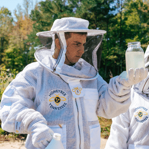Two students in beekeeping suits inspect liquid in a glass jar.