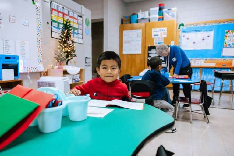 Little boy wearing a red sweatshirt is sitting behind a green table holding a pencil.