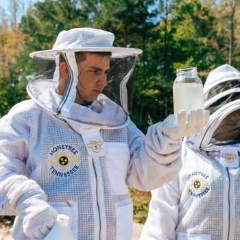 Two students in beekeeper suits inspect water in a jar.