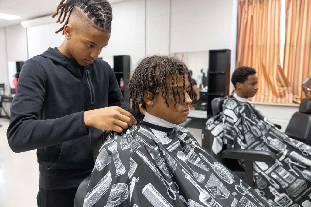 Two students sit in barber chairs with smock, one student stands behind them.
