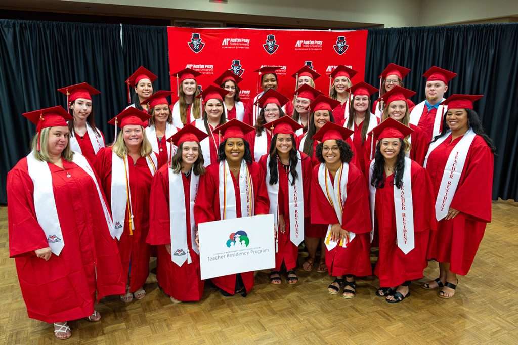 Group Photo of Graduates in red cap and gowns with Teacher Residency Program logo on poster held in front of the group