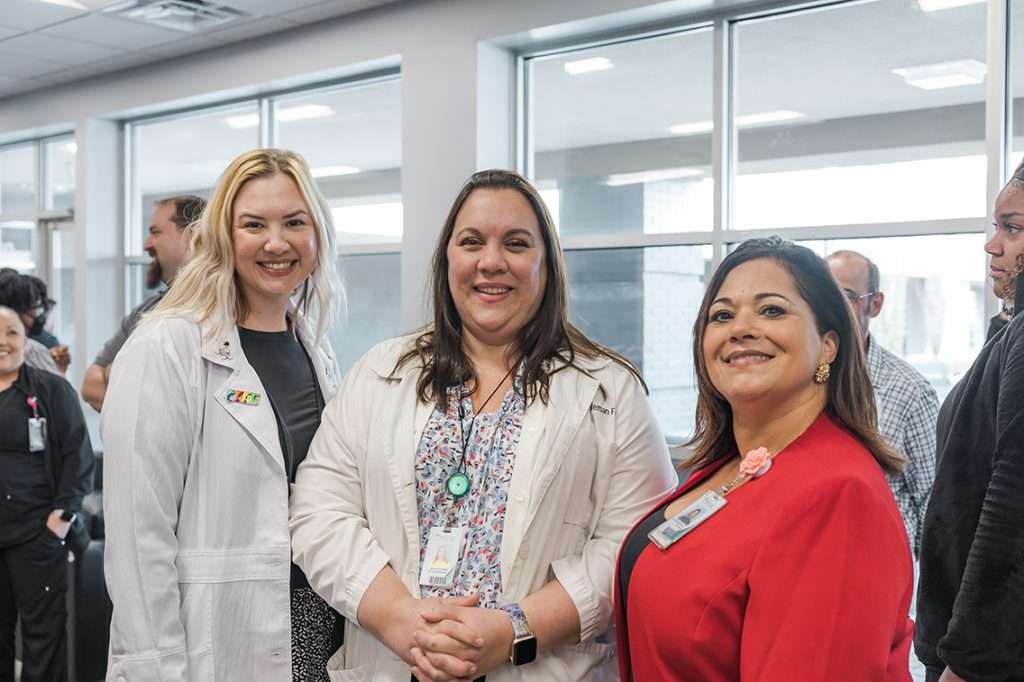 Three women are smiling in a brightly lit room with large windows.