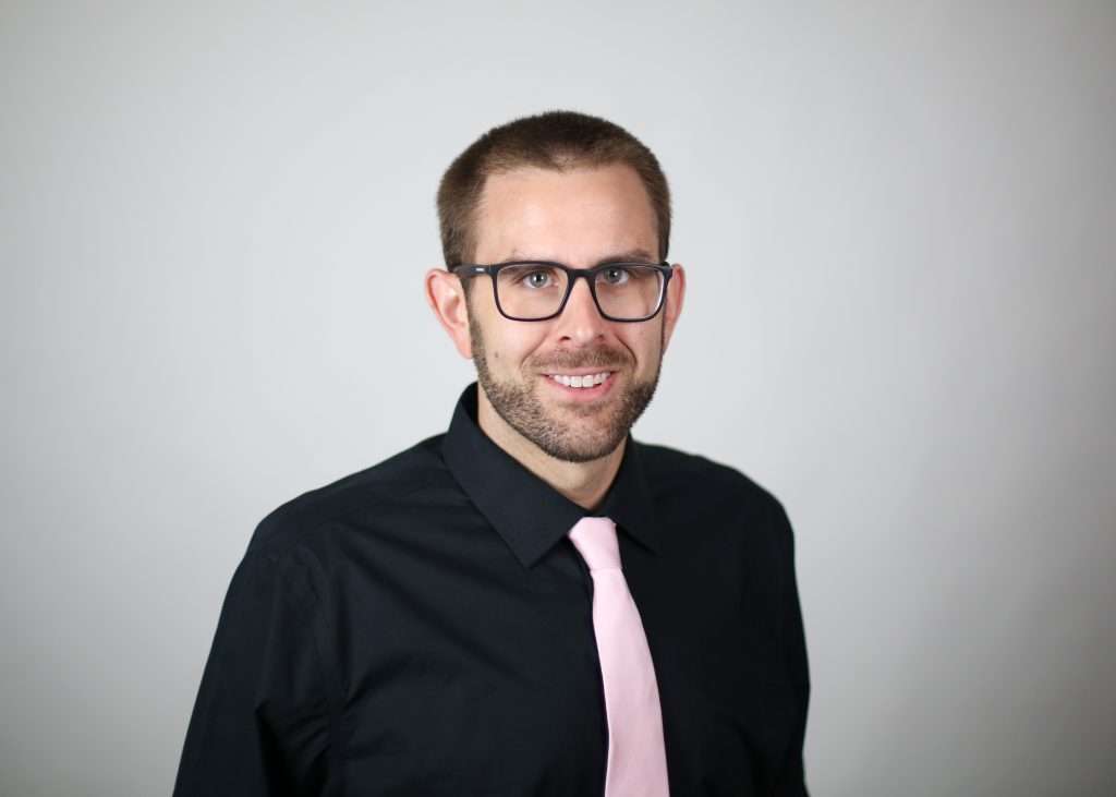 White man with glasses wearing a black shirt and pink tie. 