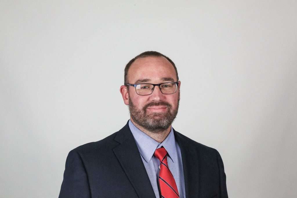 White man with glasses wearing a blazer and red tie.
