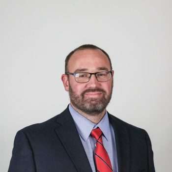 White man with glasses wearing a blazer and red tie.