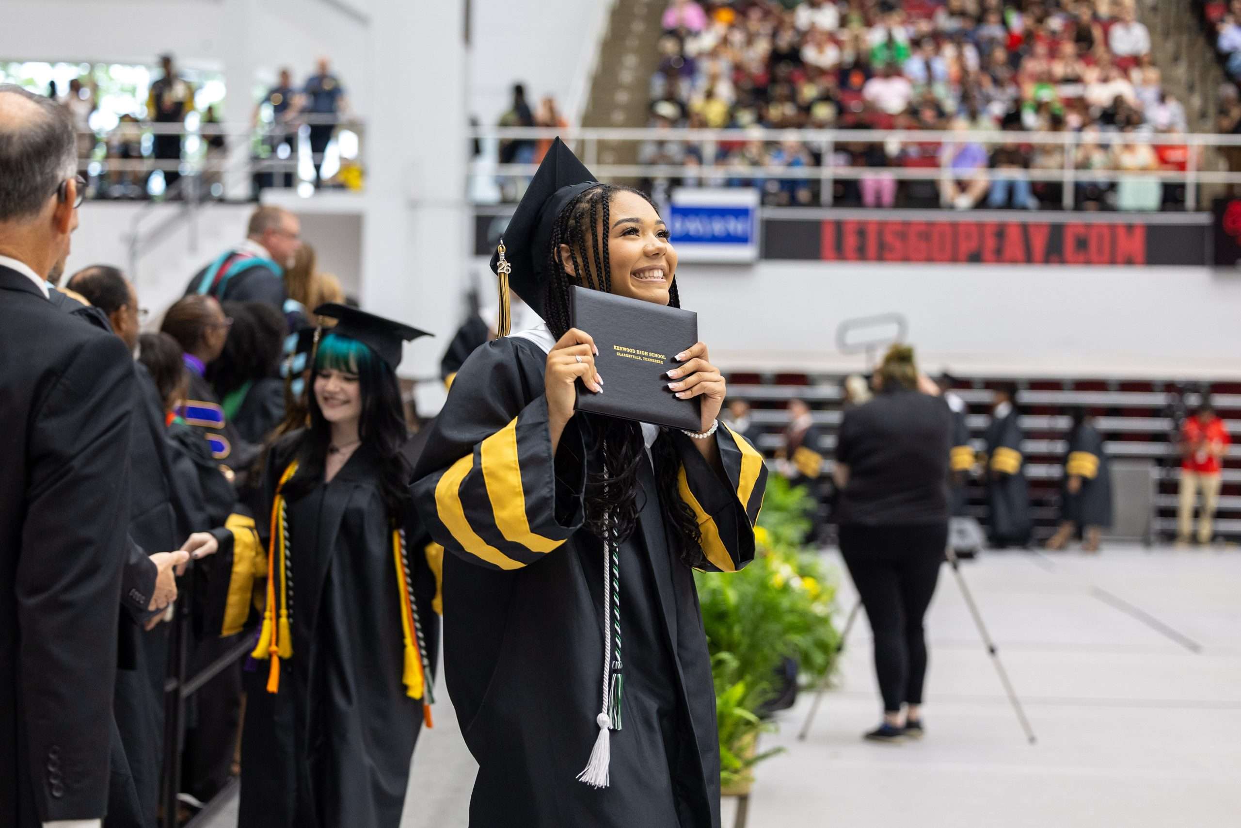 A graduate in a maroon cap and gown looks upwards, smiling amid a large, cheering crowd in an indoor arena. The atmosphere is celebratory and joyful.