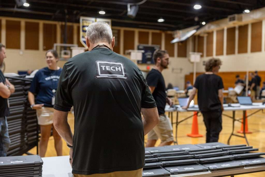 A gymnasium with people engaged in a tech setup; a man wearing a shirt that says "TECH" stands at a table with laptops, while others converse nearby.