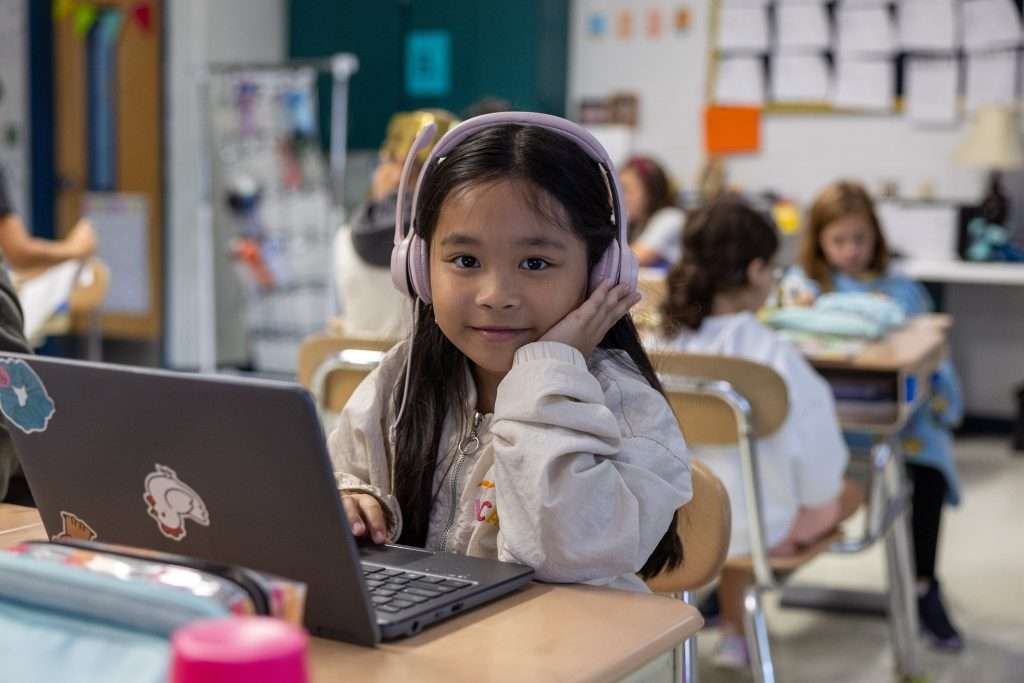 A young girl with long black hair and pink headphones sits at a desk with a laptop, looking thoughtful in a classroom filled with other students.