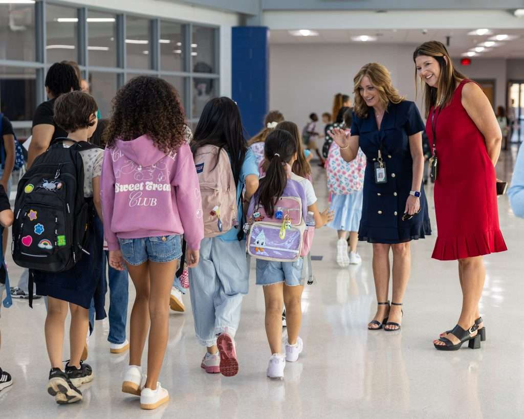 A busy school hallway with children walking away, featuring two women engaging with a girl in the foreground. One woman wears a navy dress and the other a red dress.