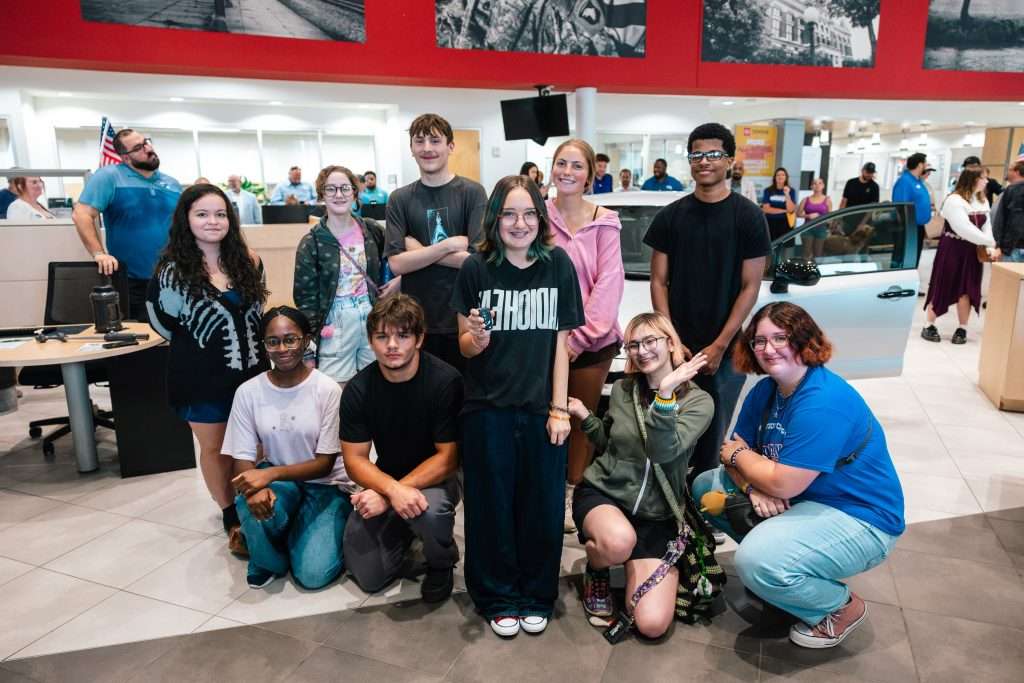 10 high school students stand in a group in front of a new car. One female student is holding the key and smiling.