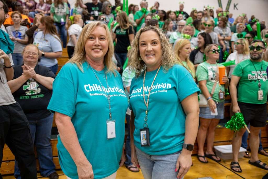 Two women in turquoise "Child Nutrition" shirts pose together in a gymnasium filled with a cheering crowd in green outfits.