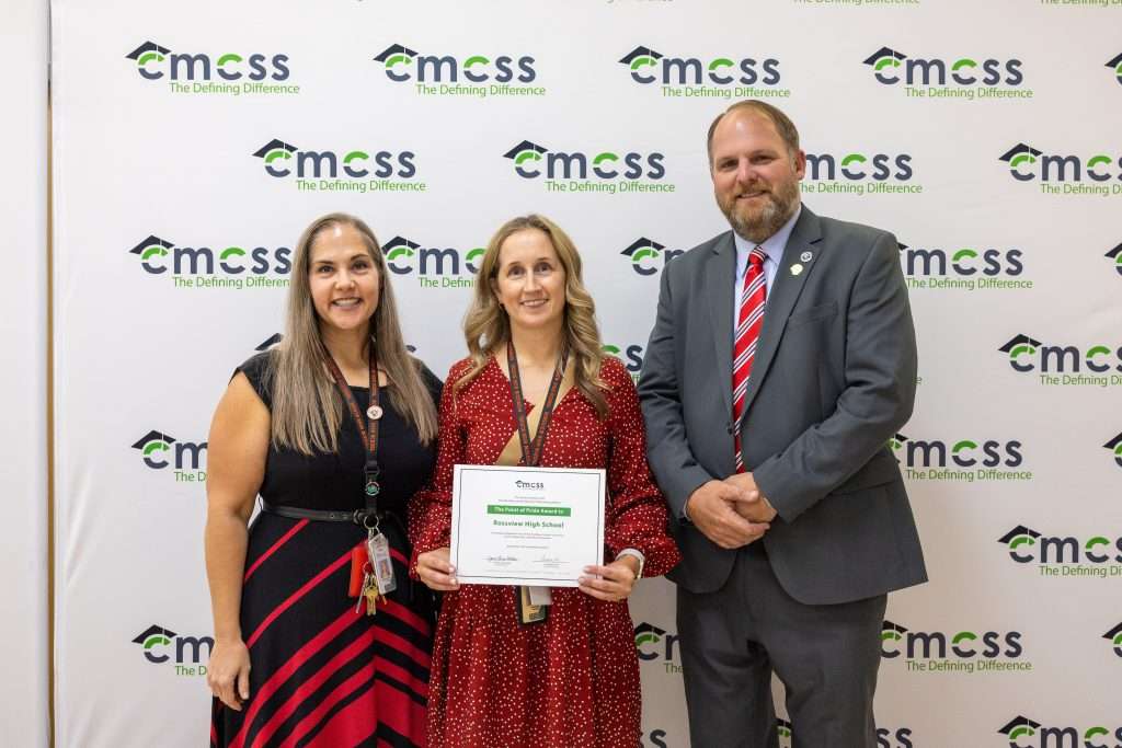 Three people standing in front of a backdrop that says CMCSS holding an award and smiling.