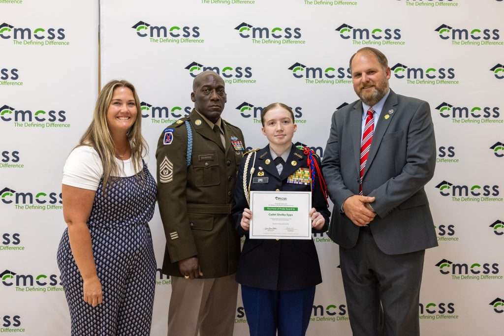 Four people standing in front of a backdrop that says CMCSS holding an award and smiling.
