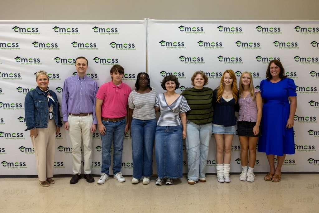 A large group of people standing in front of a backdrop that says CMCSS holding an award and smiling.