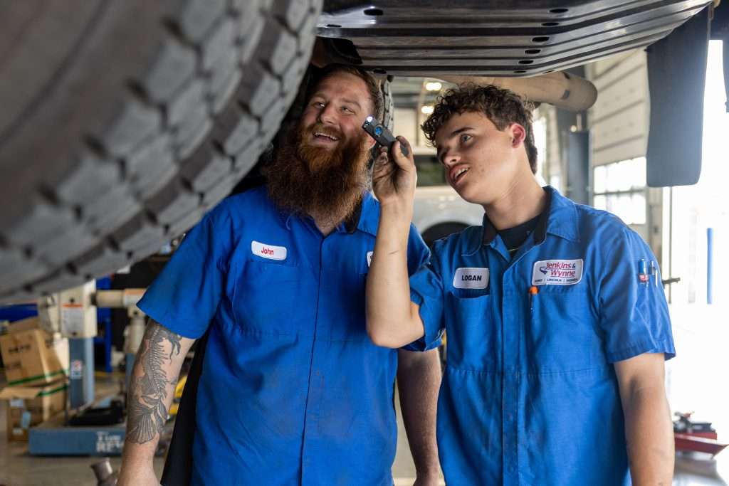 Male intern inspecting the bottom of a car with an adult male smiling