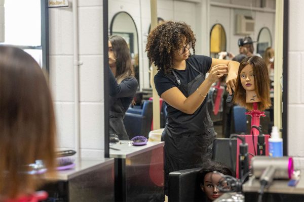 Young woman with curly hair stands in salon, styling a mannequin head with a comb. She appears focused. Mirrors and hairstyling tools are visible.